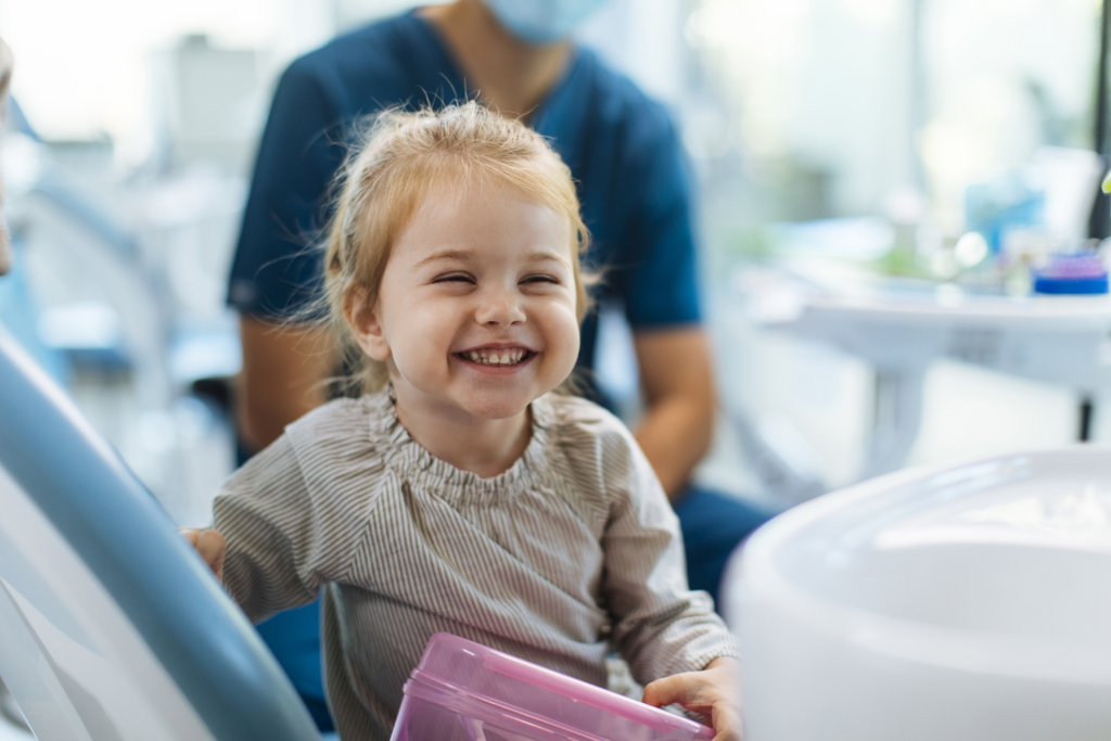 Smiling young child sitting comfortably in a dental chair during a friendly pediatric dental checkup with a dental professional nearby.