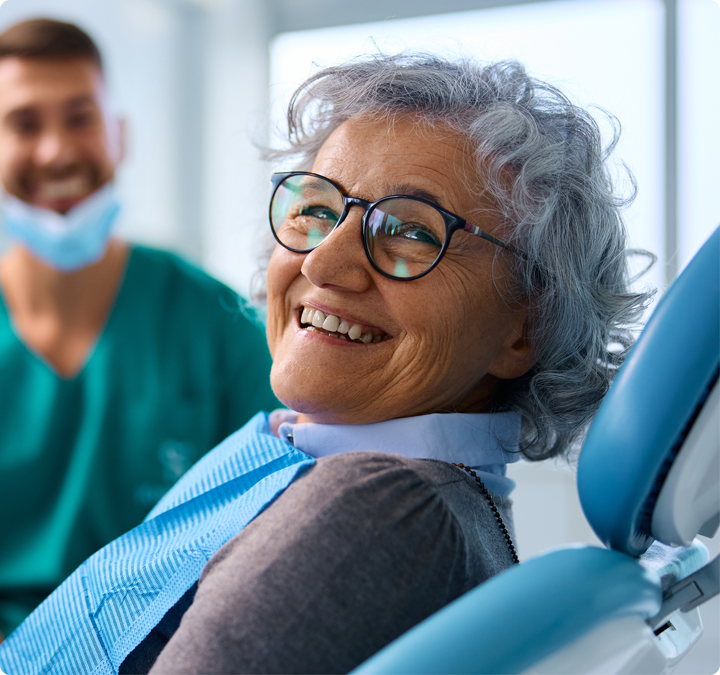 A smiling older patient sits comfortably in a dental chair while a dental professional stands nearby.